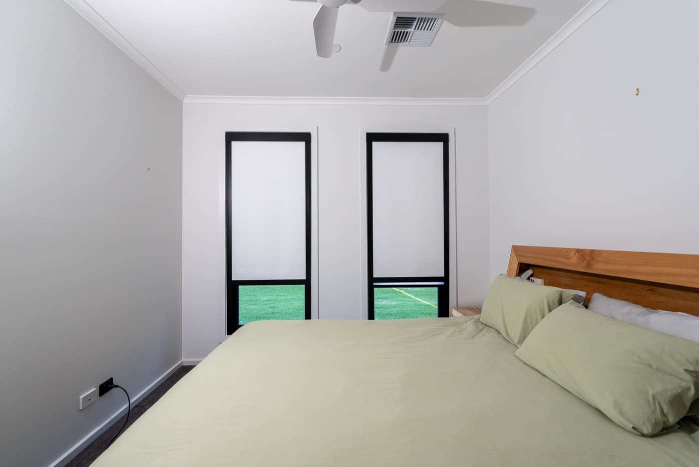 Black-frame blackout blinds lowered over two bedroom windows, sage-green bedding.