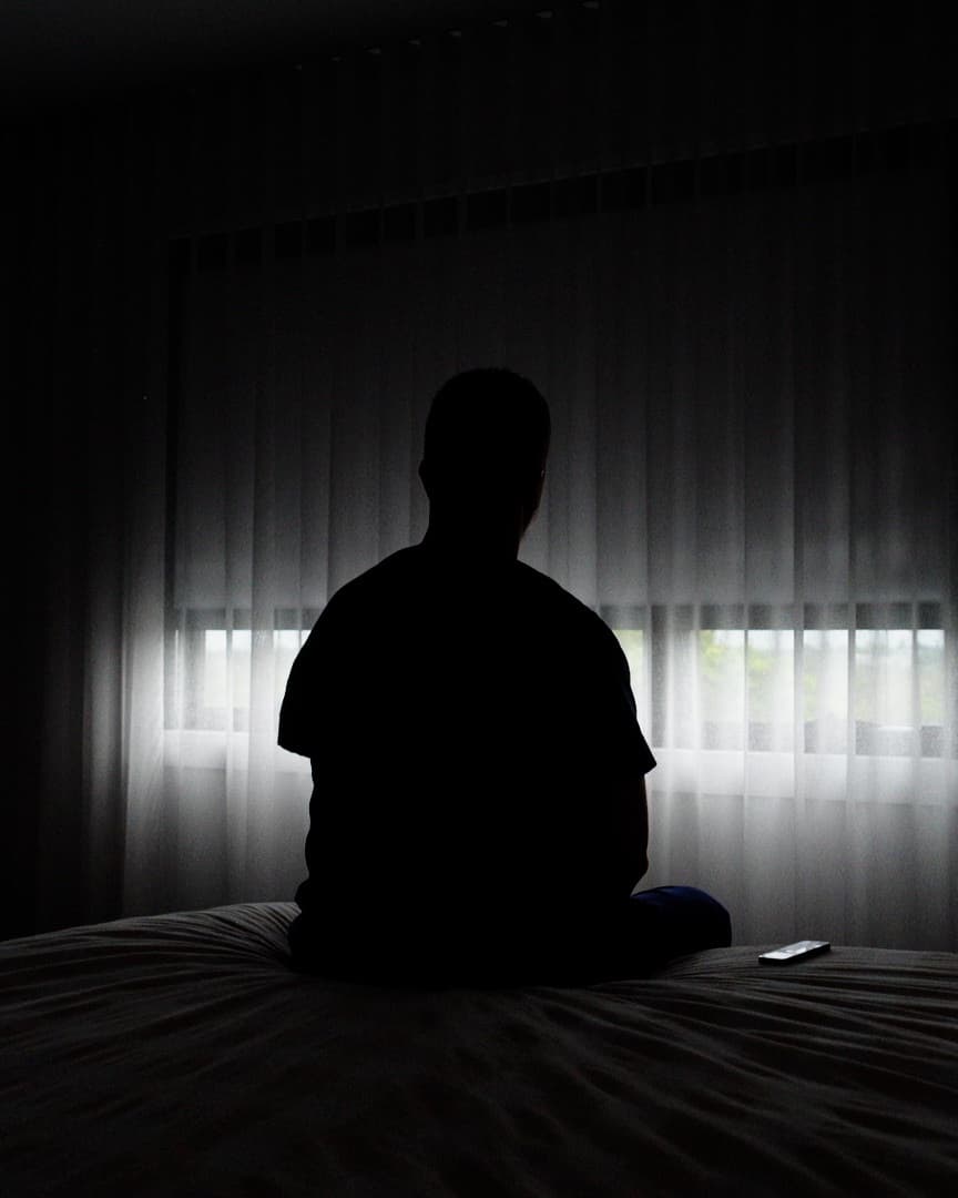 A bedroom kept dark and restful during the day, a person sitting calmly on the bed.
