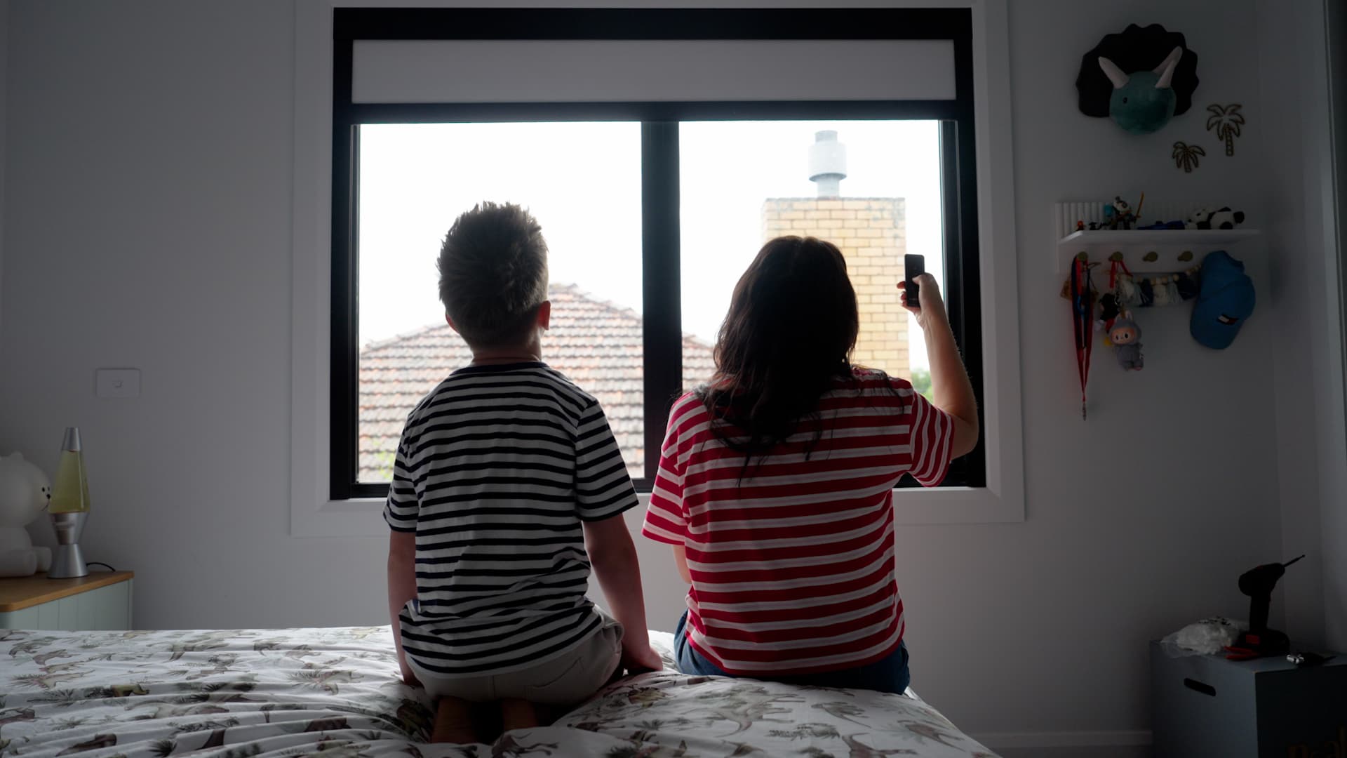 Parent and child with the remote at a black-frame bedroom window.