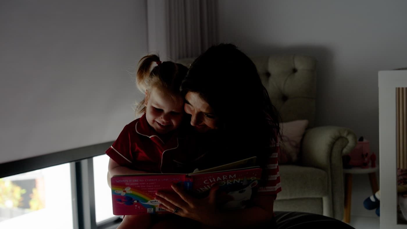 Parent reading to a child in a dim bedroom with the blind down — the calm a SmartHub schedule helps create.