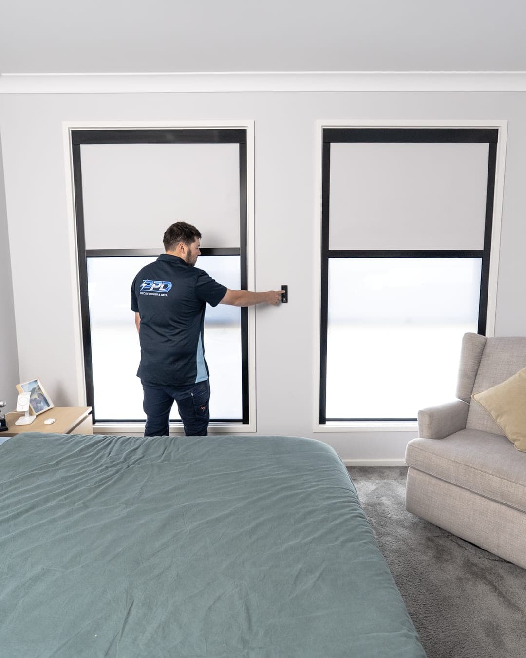 Two black-frame dual blinds in a bedroom being set from the wall remote, blackout part-down with the sheer below.