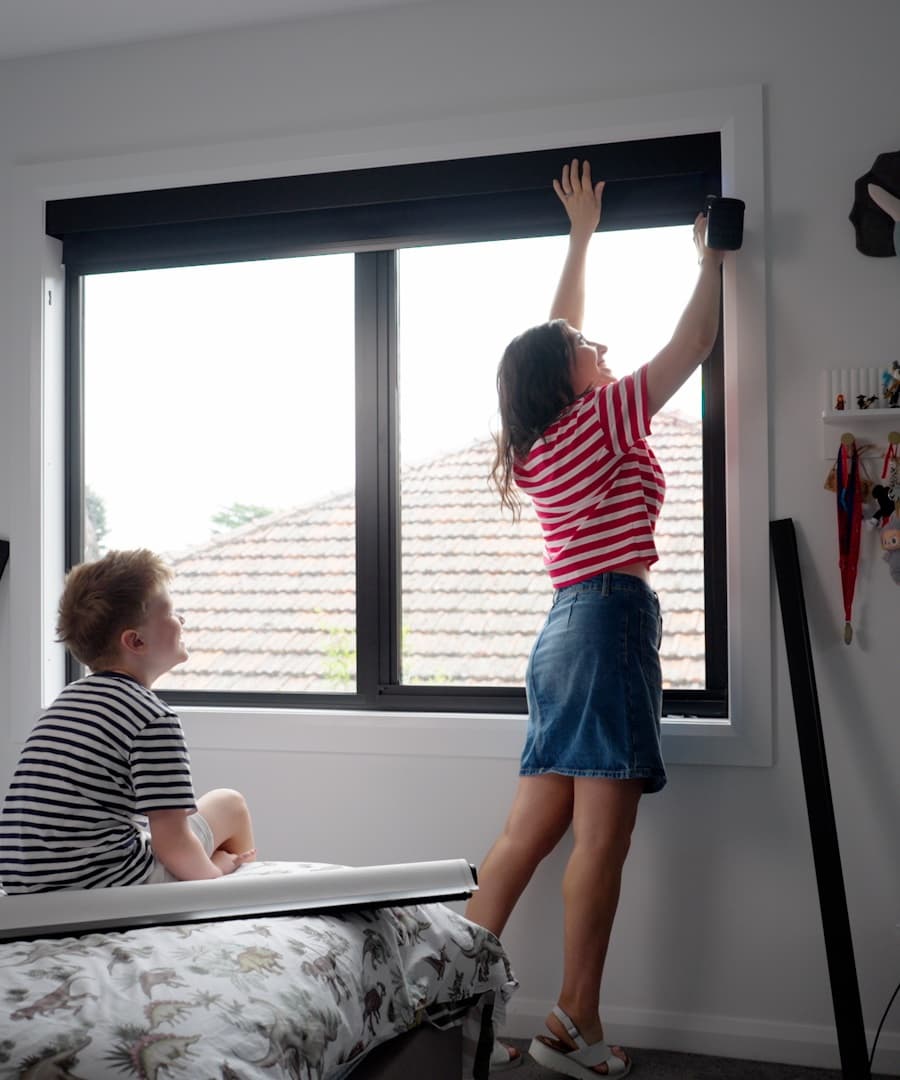 A parent fitting a motorised blackout blind at a child's bedroom window while a child watches from the bed.